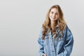 Studio portrait of pleased attractive blonde girlfriend with curls, wearing stylish denim jacket, holding hands in