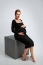 Studio portrait for pregnant women of a gorgeous young woman posing on a gray background. A woman sits on a cube and gently holds Royalty Free Stock Photo