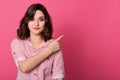 Studio portrait of pretty european female with dark hair, wearing striped blouse, pointing aside with her index finger, has calm