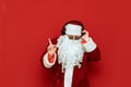 Studio portrait of Santa Claus in sunglasses listening to music in headphones and dancing on a red background. Santa party-goer in