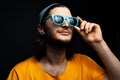 Studio portrait of smiling young man using wireless earphones wearing orange shirt, cyan sunglasses and grey hat on black. Royalty Free Stock Photo