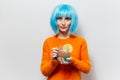 Studio portrait of young beautiful girl holding glass mug with juice and piece of lemon. Wearing blue wig and orange sweater.