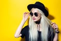 Studio portrait of young beauty blonde girl, wearing black shirt, round sunglasses, choker and hat on yellow background. Royalty Free Stock Photo
