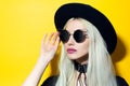 Studio portrait of young beauty blonde girl, wearing black shirt, round sunglasses, choker and hat on yellow background. Royalty Free Stock Photo