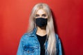 Studio portrait of young blonde girl with blue eyes in denim jacket wearing black medical face mask against coronavirus on red.