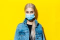 Studio portrait of young blonde girl in denim jacket wearing medical face mask against coronavirus on yellow background. Royalty Free Stock Photo