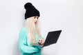 Studio portrait of young blonde girl using laptop on white background. Wearing black hat and cyan sweater. Royalty Free Stock Photo