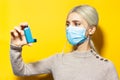 Studio portrait of young blonde girl, wearing medical flu mask and beige sweater, holding asthmatic inhaler in hand on yellow. Royalty Free Stock Photo