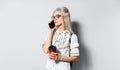 Studio portrait of young blonde teenager girl with paper cup of coffee takeaway in hand, talking on smartphone, on white Royalty Free Stock Photo