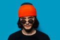 Studio portrait of young confident guy wearing sunglasses, orange beanie hat and black sweater on background of blue color. Royalty Free Stock Photo