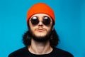 Studio portrait of young confident man on blue background. Guy with long hair wearing orange hat, round sunglasses. Royalty Free Stock Photo