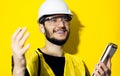 Studio portrait of young confident man, builder engineer holding notebook, using wireless earphones, wearing safety helmet.