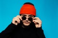 Studio portrait of young confident man wearing round sunglasses and orange beanie hat on background of blue color. Royalty Free Stock Photo