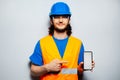 Studio portrait of young construction worker engineer wearing safety equipment, pointing finger at smartphone with mockup. Royalty Free Stock Photo