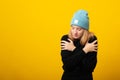 Young cute blonde girl embracing herself against yellow background, wearing blue hat and black sweater. Copy space Royalty Free Stock Photo