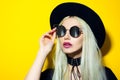 Studio portrait of young fashion blonde hipster girl with pink lips, wearing sunglasses, black hat and choker, isolated on yellow. Royalty Free Stock Photo