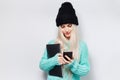 Studio portrait of young girl holding laptop, using smartphone against white background. Wearing cyan sweater and black hat. Royalty Free Stock Photo