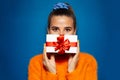 Studio portrait of young girl holding white gift box with red bow on background of blue color. Wearing orange sweater. Christmas. Royalty Free Stock Photo