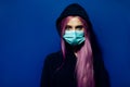 Studio portrait of young girl with pink hair and blue eyes, wearing medical flu mask and hooded sweater.