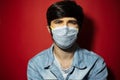 Studio portrait of young guy in denim jacket wearing medical face mask against coronavirus on red background. Royalty Free Stock Photo