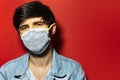 Studio portrait of young guy in denim jacket wearing medical face mask against coronavirus  on red background. Royalty Free Stock Photo