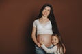 Cute little girl daughter hugging belly of her pregnant mother while posing on brown background Royalty Free Stock Photo