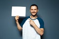 Studio portrait of young happy man holding white empty paper board with pointed finger, on background of blue color