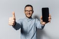 Studio portrait of young happy man showing smartphone and thumbs up on white background. Wearing eyeglasses and blue shirt Royalty Free Stock Photo