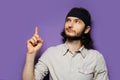 Studio portrait of young interested guy, with long brown hair, pointing forefinger up. On the background of purple wall.