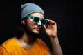 Studio portrait of young man using wireless earphones wearing yellow shirt, blue sunglasses and grey hat on background of black. Royalty Free Stock Photo