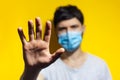 Studio portrait of young man wearing medical flu mask on background of yellow color. Close-up of male hand showing stop gesture. Royalty Free Stock Photo