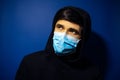 Studio portrait of young man, wearing medical flu mask and hooded sweater on background of dark blue color. Royalty Free Stock Photo