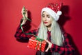 Studio portrait of young modern blonde girl, wearing Santa hat with snowflakes, trying to open a Christmas present with green bow,