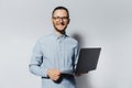 Studio portrait of young smiling man holding a laptop in hands, wearing blue shirt and eyeglasses on white background Royalty Free Stock Photo