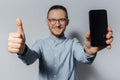 Studio portrait of young smiling man, showing thumb up and smartphone close to the camera, wearing eyeglasses and blue t-shirt on Royalty Free Stock Photo