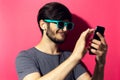 Studio portrait of young smiling man using smartphone and wireless earphones, wearing cyan sunglasses, isolated on pink coral back Royalty Free Stock Photo