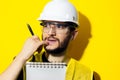 Studio portrait of young thinking man, builder engineer wearing safety helmet and glasses for construction on yellow background. Royalty Free Stock Photo