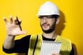 Studio portrait of a young thinking man, builder engineer wearing safety helmet and glasses for construction on yellow background. Royalty Free Stock Photo