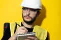 Studio portrait of a young thinking man, builder engineer wearing safety helmet and glasses for construction on yellow background. Royalty Free Stock Photo