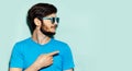 Studio profile portrait of young guy, wearing cyan sunglasses and blue shirt, pointing with finger on empty background of cyan.
