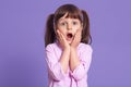 Studio shot of astonished female kid with widely opened mouth, keeping hands on cheeks, looks at camera with big eyes, standing