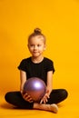 Studio shot of attractive little gymnast girl blonde wearing black legends and a bathing suit with a purple ball Royalty Free Stock Photo