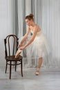 Studio shot of a calm beautiful ballerina in a white airy classic dress put her foot on a vienna chair and tying ribbons on pink