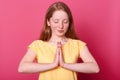 Studio shot of calm beautiful Caucasian teenager girl stands isolated over pink background while praying, keeps eyes closed, asks Royalty Free Stock Photo