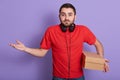 Studio shot of caucasian bearded man dressed red casual t hirt standing isolated over lilac background with carton box in hand,