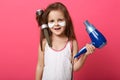 Studio shot of charming cute female kid posing isolated over rose studio background with blue hairdryer in hands, little girl with