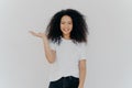 Studio shot of cheerful African American woman grins happily, keeps palm raised over copy space, pretends holding something, wears Royalty Free Stock Photo