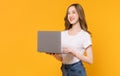 Studio shot of cheerful beautiful Asian woman in white t-shirt and holding laptop with typing on keyboard on yellow background