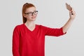Studio shot of cheerful red haired female in eyewear, makes selfie on cell phone, poses in camera of digital electronic device, st