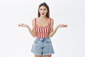 Studio shot of concerned questioned and frustrated displeased woman in striped top and denim shorts shrugging with hands